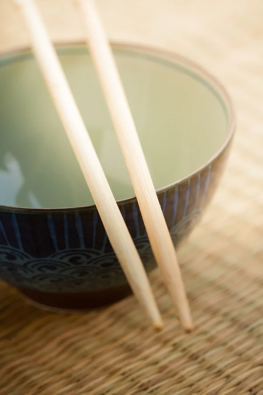 A pair of wooden chopsticks resting across a ceramic bowl with a blue wave pattern, placed on a woven bamboo mat.